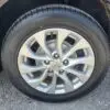 Close-up of a 2017 Nissan Sentra SV wheel with a silver alloy rim, black tire, and circular design featuring multiple spokes. The central badge shows the Nissan logo. The car is parked on gravel.