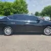 A 2017 Nissan Sentra SV sits on a gravel lot. The black sedan boasts tinted windows and shiny alloy wheels. In the background, other vehicles, trees, and a partly cloudy sky are visible.