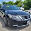 A 2015 Honda Accord Touring V6 in black is parked in a sunny lot, with a yellow Loaded Navigation sign on the windshield. The scene features trees and other cars, enhancing the picturesque backdrop for this sleek sedan.