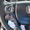 Close-up of a 2015 Honda Accord Touring V6 steering wheel with various control buttons on the left, like volume, phone, and source. The centered Honda logo is on textured black leather with visible airbag markings.