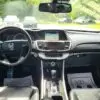 The 2015 Honda Accord Touring V6s interior features a steering wheel, dashboard with touchscreen, and gear shift. Sunlight highlights the black leather seats and two thank-you mats on the floor through the front windshield.