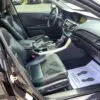 The 2015 Honda Accord Touring V6s interior includes black leather front seats, a sleek center console, steering wheel, and dashboard. Sunlight illuminates the paper floor mat with shoe prints on the passenger side floor as the inviting door stands open.