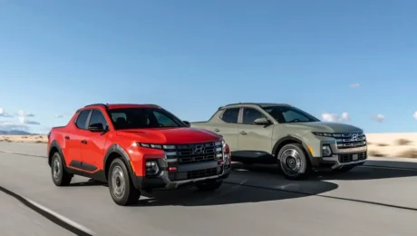 Two Hyundai Santa Cruz pickup trucks cruise along a desert road. The truck on the left is a refreshed red, while the one on the right is a sleek gray. With clear skies above and dry, sandy terrain below, they embody adventure despite any price hike.
