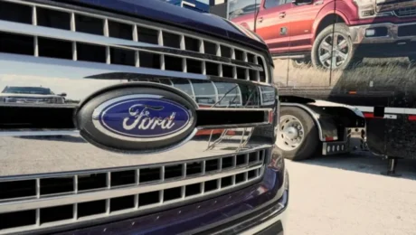 Close-up of a Ford trucks front grille boasting the iconic blue and silver logo. In the background, another truck is partially visible with an image of a red pickup on its side. The scene unfolds outdoors on a sunny day, reflecting Fords enduring commitment amidst conservative pressure.