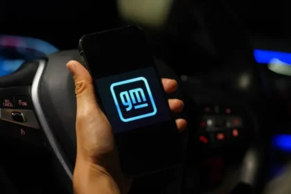 A hand holds a smartphone displaying the GM logo in front of a car steering wheel. The dimly lit car interior features visible controls and ambient blue lighting, hinting at the allure of modern design despite past concerns like faulty transmissions.