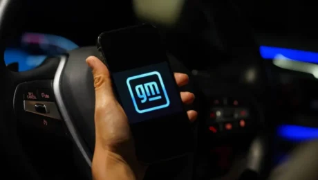 A hand holds a smartphone displaying the GM logo in front of a car steering wheel. The dimly lit car interior features visible controls and ambient blue lighting, hinting at the allure of modern design despite past concerns like faulty transmissions.