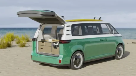 A green and white ID.Buzz van, reminiscent of classic Peace Vans, is parked on a sandy beach with its rear door open. A surfboard rests atop the roof, while inside reveals a compact kitchen setup. The ocean and some coastal vegetation stretch under the cloudy 2025 sky.