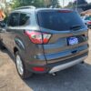A gray 2018 Ford Escape SEL 4WD sits on gravel in a car lot, with a Loaded sign displayed. Surrounding it are trees and various vehicles beneath a clear sky.
