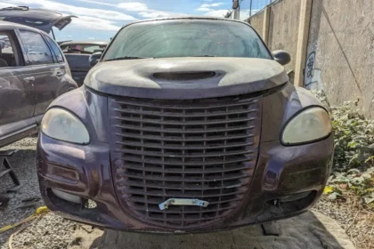 Front view of an old, dusty, dark purple Chrysler PT Cruiser with a large grille, parked in a lot. This 2002 model features a hood scoop and foggy headlights. Another limited edition car is partially visible to the left. The scene unfolds outdoors on a sunny day.