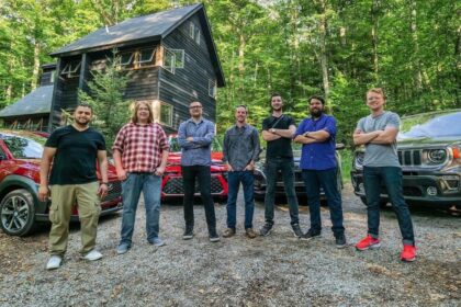 Seven people stand in a gravel driveway before a dark wooden house nestled among trees. Behind them, three cars—a red car on the left, one in the middle, and a green SUV on the right—reflect 20 years of evolution in the auto industry. Seven people stand in a gravel driveway before a dark wooden house nestled among trees. Behind them, three cars—a red car on the left, one in the middle, and a green SUV on the right—reflect 20 years of evolution in the auto industry.