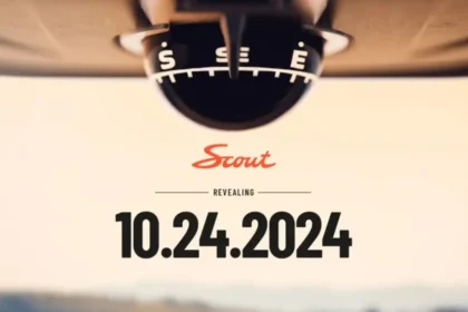 Close-up of a compass with S and SE visible at the top, below which Scout appears in red script. The word Revealing is prominently displayed above the official reveal date, 10.24.2024, hinting at a new era of EVs against a blurred background.
