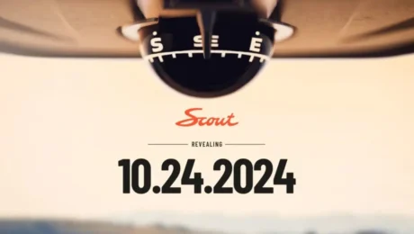 Close-up of a compass with S and SE visible at the top, below which Scout appears in red script. The word Revealing is prominently displayed above the official reveal date, 10.24.2024, hinting at a new era of EVs against a blurred background.