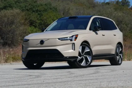 A beige Volvo EX90 SUV is parked on a paved outdoor area with greenery in the background. It boasts sleek, modern design features, including distinctive headlights and alloy wheels. This luxury electric vehicle is well-lit under a clear sky.