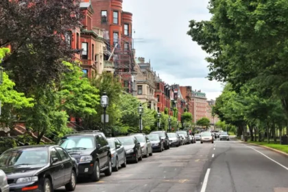 A tree-lined urban street with convenient parking on one side. A row of historic brownstone buildings with green staircases aligns the street. The sky is overcast, and vintage street lamps are visible along the sidewalk.