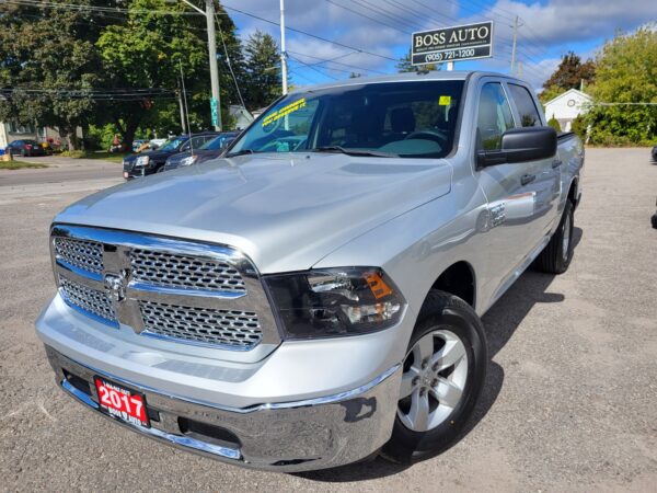 A 2017 Dodge Ram 1500 ST 4X4 with a silver finish and chrome grille is parked near Boss Auto dealership. Its license plate confirms its vintage, set against a backdrop of blue sky and trees, highlighting this robust pickup truck.