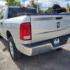 A silver 2017 Dodge Ram 1500 ST 4X4, adorned with a 4x4 Boss & Life plate on the hitch, is parked on sunlit asphalt. Trees and houses provide a serene backdrop, accentuating the rugged presence of this truck.