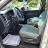 Interior of a 2017 Dodge Ram 1500 ST 4X4 with gray seats and black dashboard. The drivers seat has paper on the floor, the center console is between front seats, and logo is on steering wheel. Houses and greenery are visible through windows in this rugged pickup truck.