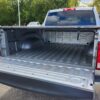 The open tailgate of a 2017 Dodge Ram 1500 ST 4X4 shows its spacious, empty bed. This silver pickup truck is parked on gravel, with trees and other vehicles subtly in the background.