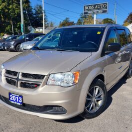 A Boss Auto dealership features a beige 2013 Dodge Grand Caravan SXT w/ DVD. Parked on a sunny day with its year on the license plate, it is surrounded by other cars and lush trees, suggesting an ideal setting for an adventurous road trip.