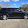 A sleek 2017 Dodge Journey SE sits on a gravel lot surrounded by trees under a blue sky with scattered clouds, while other vehicles in the background highlight the SUVs robust black design.