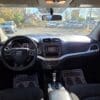 Inside the 2017 Dodge Journey SE, sunlight illuminates the front seats and mats reading YOUR BUSINESS IS APPRECIATED! The view includes the steering wheel with the Dodge logo, a central dashboard screen, gear shift, and visible cars outside.
