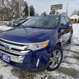 A blue 2013 Ford Edge SEL V6 SUV with chrome accents is parked on a snowy lot in front of Boss Auto, reflecting the wintry scene, while other cars and bare trees form the backdrop.