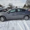 A gray 2018 Nissan Sentra SV is parked in a snowy lot amid other vehicles. The ground is blanketed in snow, with bare trees and suburban houses in the background. A cloudy sky looms above, enhancing the wintery chill.