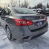 A gray 2018 Nissan Sentra SV sits on a snowy street with a Gas Saver sign highlighting its efficiency. Other cars and trees are visible in the background under a cloudy sky, enhancing the serene winter atmosphere.