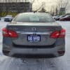 Rear view of a 2018 Nissan Sentra SV in gray with a GAS SAVER dealer plate, parked on a snowy lot with trees, other cars, and a yellow building in the background.