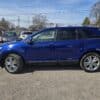 A 2013 Ford Edge SEL V6 SUV in dark blue is parked on a gravel lot under a clear sky. Several other vehicles are visible in the background, with trees and utility poles in the distance.
