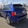 A 2013 Ford Edge SEL V6 in blue is parked on a paved area under a clear sky, with the rear and side visible. A Certified sign is on the license plate area, and trees and utility poles decorate the background.