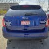 Back view of a 2013 Ford Edge SEL V6 in blue with a certified pre-owned plate from Boss Auto, basking in sunlight amid other cars and leafless trees on this bright day.