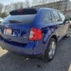 A blue 2013 Ford Edge SEL V6 SUV is parked in an outdoor lot with a sunny backdrop. The vehicles rear and side are visible, along with other cars and bare trees in the distance.