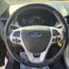 Close-up of the 2013 Ford Edge SEL V6 steering wheel with the Ford logo, a dashboard showcasing gauges and a screen. Sunlight illuminates the interior through the windshield.