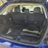 A 2013 Ford Edge SEL V6 with a blue exterior is shown with its trunk open. Car mats are neatly laid flat, and the rear seats remain upright, offering a glimpse of its spacious design. Other cars and trees are visible in the background, emphasizing the SUVs practicality and elegance.