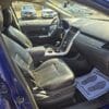 Inside the 2013 Ford Edge SEL V6 in blue, you see black leather seats, a central console with cup holders, and a dashboard. A paper mat on the passenger floor reads Your business is appreciated, while other cars are visible through the windows.