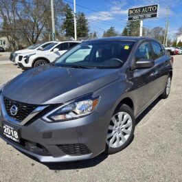 A gray 2018 Nissan Sentra SV is parked at a dealership lot under clear skies, with other cars and a Boss Auto sign in the background.