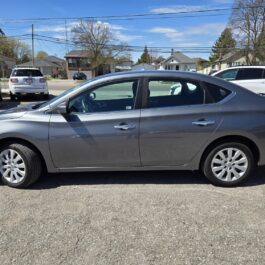 Side view of the 2018 Nissan Sentra SV parked in a lot on a sunny day, with other cars and houses visible in the background.