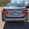 A 2018 Nissan Sentra SV in gray is parked outdoors on a sunny day, seen from the rear with a Gas Saver license plate frame from Boss Auto. Other cars and bare trees appear in the background.