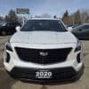 Front view of a white 2020 Cadillac SUV displayed in a used car lot. The car has a prominent grille and is surrounded by other parked vehicles. A sign above reads Boss Auto. Trees and a clear sky are in the background.