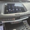 Close-up of a car dashboard showing an infotainment screen with icons for audio, phone, settings, Apple CarPlay, and Android Auto. Below are buttons for climate control and hazard lights. Parts of the steering wheel and a magazine on the floor are visible.