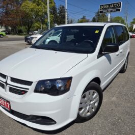 A 2017 Dodge Grand Caravan SE in white is parked at a car dealership lot on a sunny day, with other vehicles and the BOSS AUTO sign visible in the background.