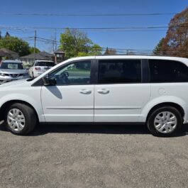 A 2017 Dodge Grand Caravan SE in white is parked in a lot on a sunny day, with other cars and houses in the background.