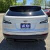 A 2020 Cadillac XT4 Sport AWD in white is shown from the rear, parked in a lot with a blue LIKE NEW dealer plate, trees, and other vehicles in the background.