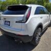 A 2020 Cadillac XT4 Sport AWD is parked outdoors on a sunny day, shown from the rear left side. The car is clean with shiny alloy wheels, and its license plate reads LIKE NEW. Trees and other cars can be seen in the background.