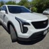 A 2020 Cadillac XT4 Sport AWD SUV in white is parked on a paved lot, angled slightly left, with trees and other cars visible in the background.