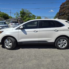 A 2017 Ford Edge SEL is parked in a lot on a sunny day, surrounded by other vehicles and trees, with houses and power lines in the background.
