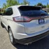 A 2017 Ford Edge SEL is parked on a sunny day in a lot. The license plate frame reads LIKE NEW, with trees, houses, and a clear blue sky in the background. This used SUV offers style and comfort.