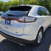 A 2017 Ford Edge SEL in white is parked outdoors on asphalt, viewed from the rear passenger side. It features a LIKE NEW dealer plate and is surrounded by trees and other used Ford SUVs.