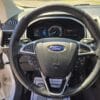 A detailed look at the 2017 Ford Edge SELs steering wheel and dashboard, highlighting control buttons, speedometer, and digital displays with sunlight streaming through the open driver-side door.